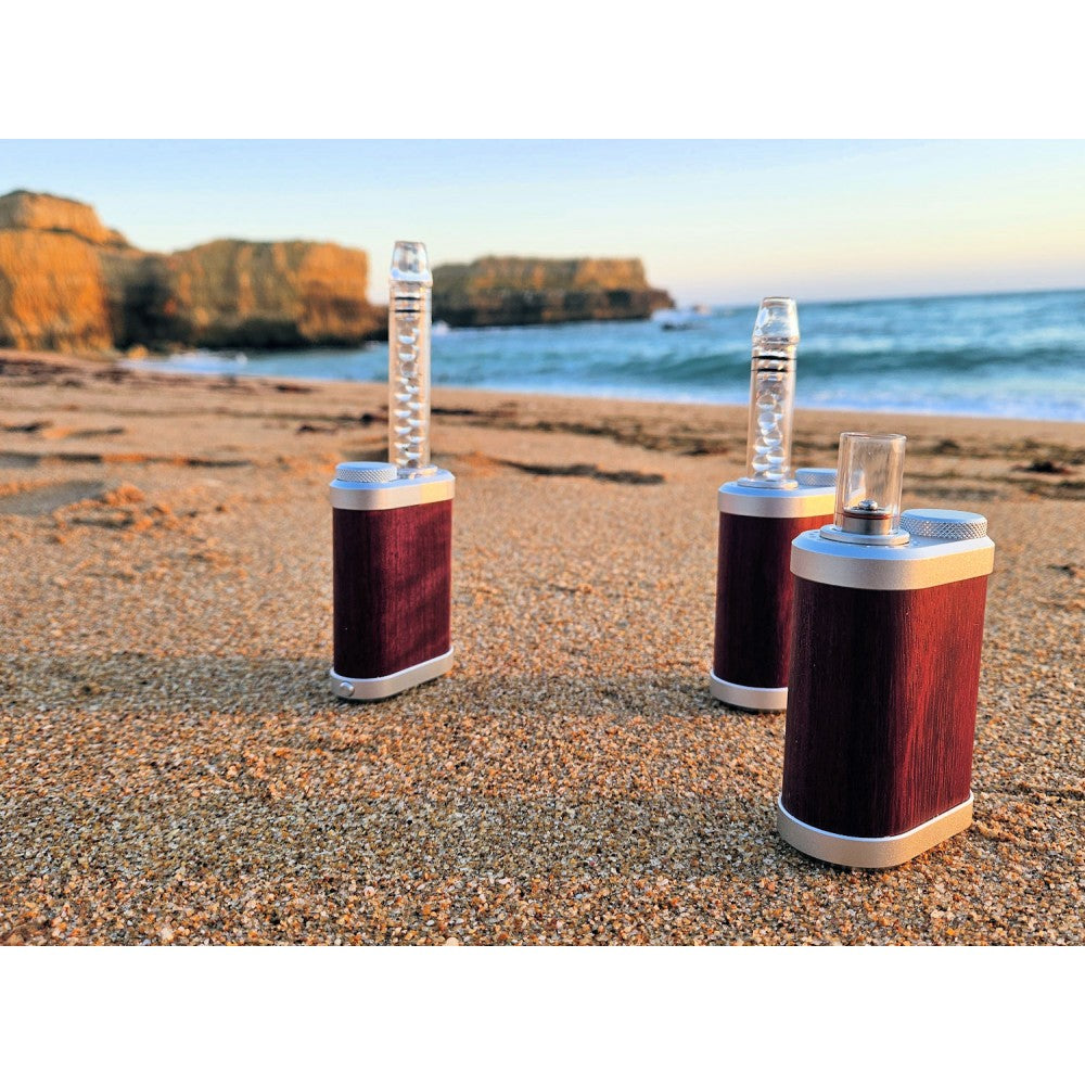 Three Tinymight 2 Purple Heart vaporizers on a sandy beach with ocean and rocks in the background